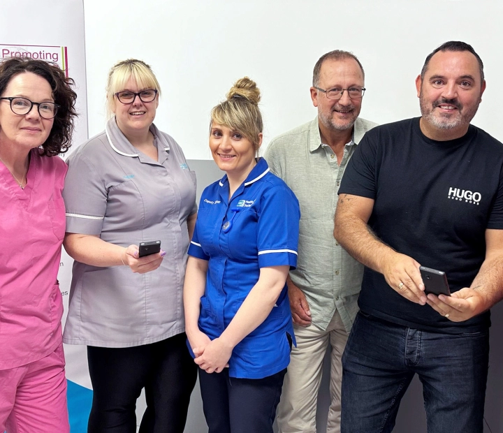 Петима усмихнати души стоят заедно в здравно облекло, като двама от тях държат преводач.