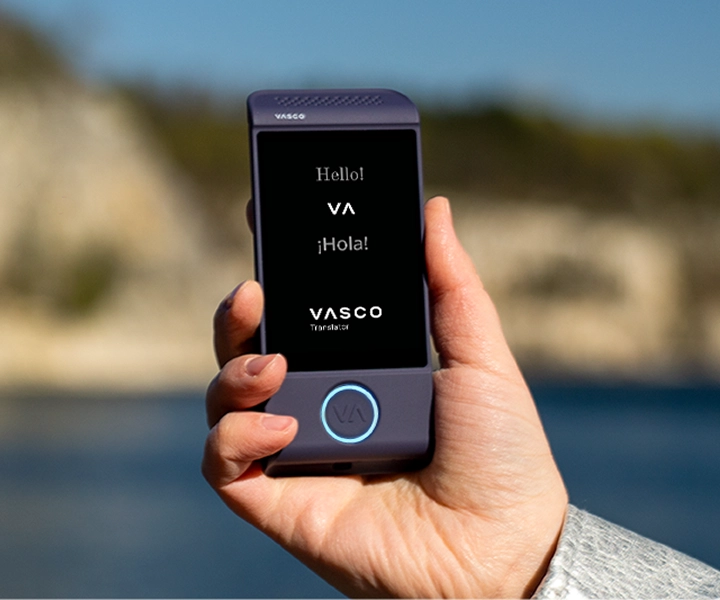 Близък план на ръка, държаща лилаво електронно устройство за превод Vasco Translator M4, на ярък външен фон. Екранът показва поздравите „Hello!“, „¡Hola!“ и логото на „VASCO Translator“.